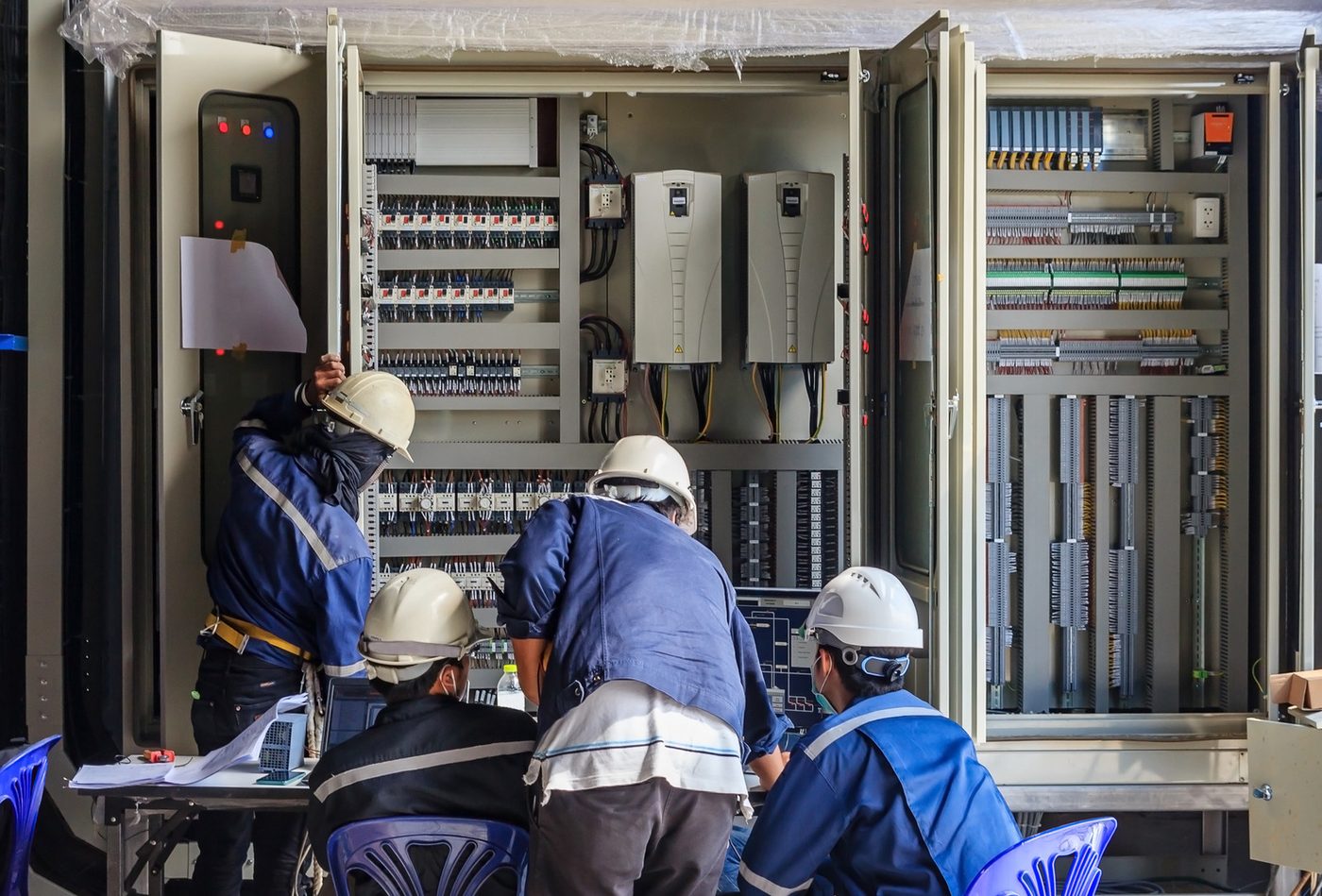 Equipe técnica com capacetes trabalhando em painéis elétricos abertos